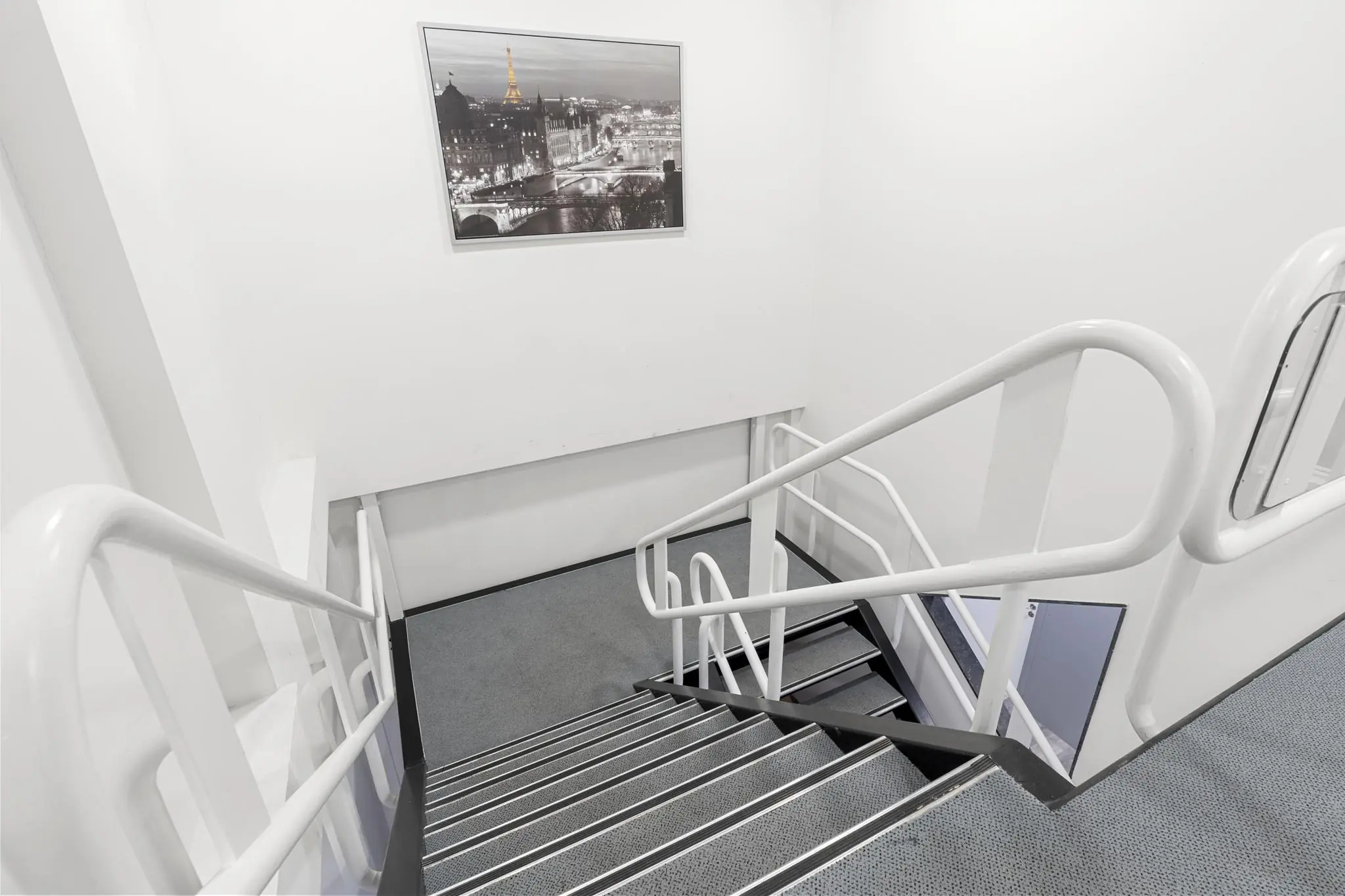 Witgeschilderd trappenhuis met grijze vloerbedekking en een ingelijste zwart-witfoto van Parijs aan de muur.